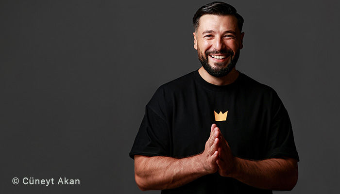 Der Comedian Cünyet Akan lacht in die Kamera. Er trägt ein T-Shirt mit einer stilisierten Krone.